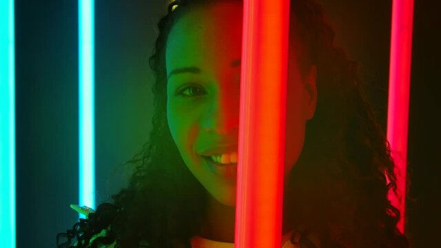 Portrait of a young lovely female African American with long curly hair poses against a dark studio with bright multicolored neon lights. Slow motion. Close up.