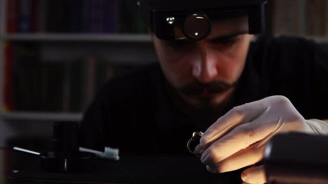 Close-up of young caucasian goldsmith who is examining and then cleaning ring by brush. Portrait of man with loupe on his head who is working in pawnshop. Concept of working in precious metal's sphere