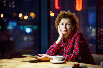 Mature caucasian woman drinking coffee and watching an electronic tablet, use internet. sitting inside city cafe at evening. Elderly female uses technology in coffee shop sit by window with city view