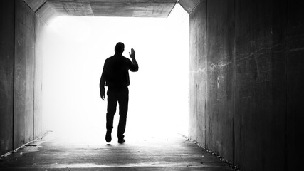 A single male in a dark tunnel exiting and walking towards a bright white light while waving goodbye or hello.