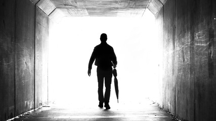 A silhouette of a man holding a folded up umbrella leaving a dark tunnel walking towards the light. Weather, climate, leaving and departure theme