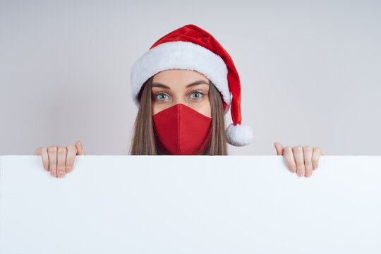 Nice woman in a red sweater, in a protective mask, in a Santa hat, holds a white banner, space for text, relama.