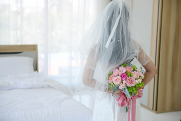 Asian bride wear the white wedding dress standing back and holding pink rose flowers at the back at the front of the bed