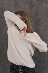 A girl in a light sweater on a gray background. Women's hands close-up. The girl covers her eyes with her hands