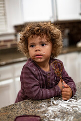 Cute mixed-race boy licking the blender beater from a bowl of brownie cake batter as he makes a mess in the kitchen. Child having fun at home baking and cooking