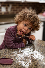 Cute mixed-race boy licking the blender beater from a bowl of brownie cake batter as he makes a mess in the kitchen. Child having fun at home baking and cooking