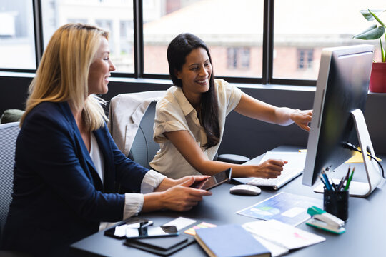 Diverse businesswomen sitting by desk using computer discussing in office