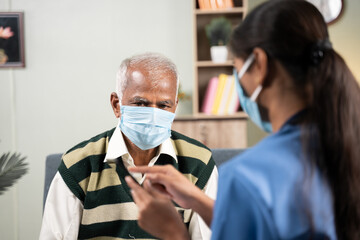 Shoulder shot of Doctor explaining how to use health app on mobile to elder man during home visit while both worn face mask - concept of senior people helath check up.