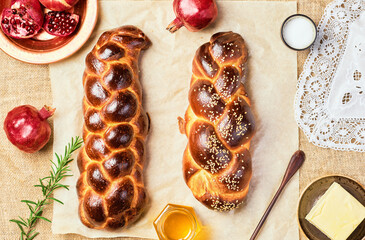 Challah bread, freshly baked sweet braided bread on parchment, sackcloth background. Pomegranates, apples, butter and honey for bread on the table. Top view, product layout, hanukkah celebration