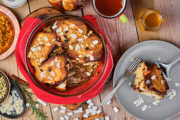 Freshly baked bread pudding with raisins, almond flakes and cardamom in a baking dish on the table. Dessert concept, pudding slice on a plate. Top view, wooden table
