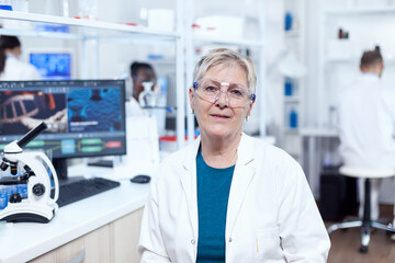 Senior medical research in scientific clinic with virus on computer display. Elderly scientist wearing lab coat working to develop a new medical vacine with african assistant in the background.