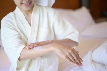 A happy beautiful woman in white bathrobe applying body moisturizing cream in bedroom, skin care and treatment concept