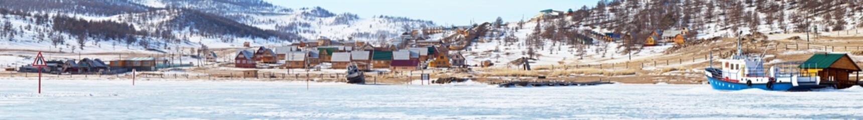 Winter Baikal Lake. A panoramic view of the coast of the Kurkut Bay - a popular tourist destination with wooden houses, a ship on a frozen pier and an exit to the ice road to Olkhon Island