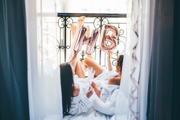  Brunette young women in white bathrobes clink elegant glasses with delicious champagne lying on...