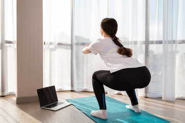 Fototapeta premium A young woman in sportswear trains at home for online lessons. Rear view. Concept of home sports training on videocommunication