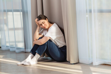 Depressed crying woman holding her head with her hand and sitting on the floor. Copy space. Windows on the background. The concept of domestic violence