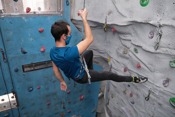 Young caucasian man wearing protective face mask rappelling at indoor artificial rock climbing wall. New normal concept.