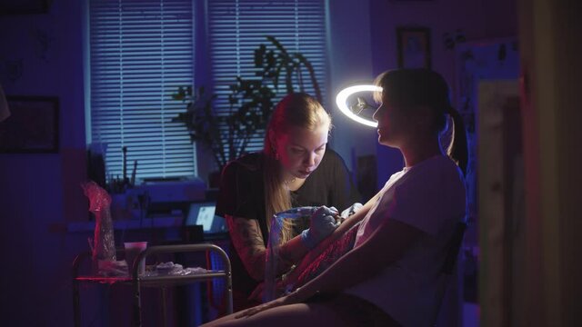 Young woman getting a tattoo on her arm by a woman master in dark neon studio