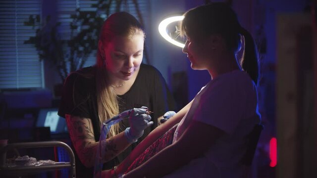 Young woman getting a tattoo by a woman master in dark neon studio