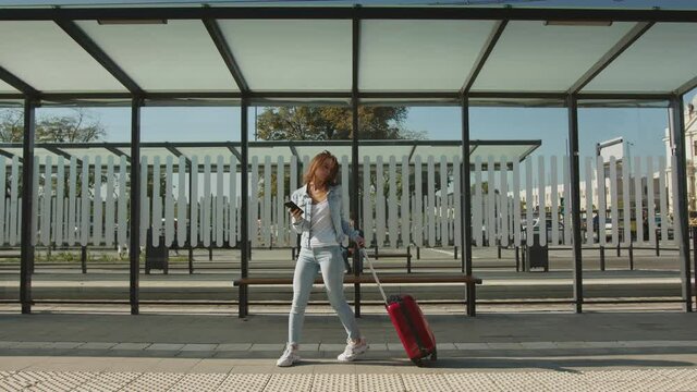 A young woman is walking through a public transport stop. She is stopping on the platform. She is holding a suitcase and texting on her smartphone. 4K.