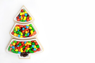 Small bright colorful candies in a hard chocolate glaze on a plate in the form of a Christmas tree on a white background.Creative idea for a pastry shop.Flatlay.Top view.