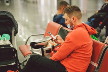 A man is waiting for his flight, sitting in an airport lounge wearing a protective mask with a phone in his hands.