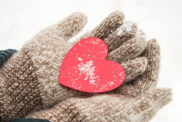 Women's hands in knitted warm wool gloves hold a red heart, against the background of snow, in winter, Valentine's day concept, close-up