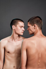 Fototapeta na wymiar Vertical shot of two young caucasian half naked twin brothers looking at each other while standing isolated over grey background