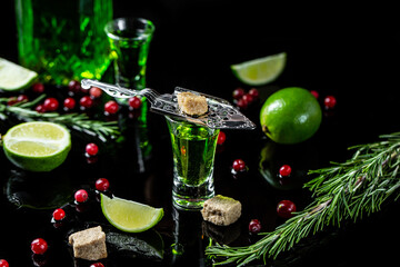 absinthe in glass with lime slices, sugar cubes, cranberries and lime, stainless steel spoon on dark background