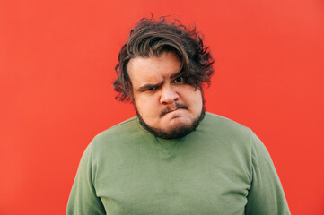 Fototapeta na wymiar Closeup portrait of an angry bearded hispanic man experiencing rage and being furious, frowning, looking at the camera with a dissatisfied gaze, standing on a red background.