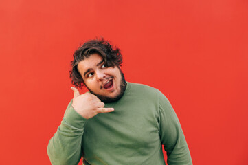 Attractive corpulent hispanic guy is showing a calling gesture, imitating a conversation, feeling cheerful and happy, standing on a red background.