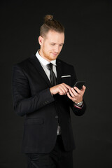 Portrait of young smart confident and successful businessman in black suit using smartphone isolated on black background