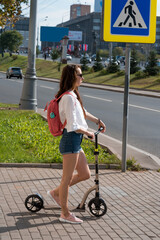 woman on scooter in summer, stands at an intersection by road, waiting to cross traffic light. Short denim shorts, white shirt and pink bag on back.