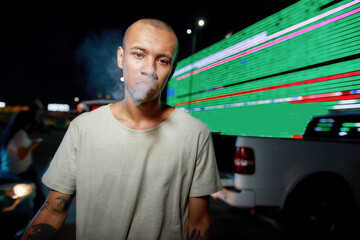 A close up of a young dark-skinned casually dressed tattooed man looking into a camera while breathing out smoke having friends and a led screen on a background