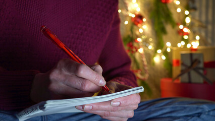 Woman in a red sweater and jeans writes a letter with a red pen to Santa Claus on the background of Christmas decor.Close-up