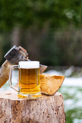 Nahaufnahme von einem Glas Bier und eine Axt auf einem Holzstock im Schnee. Winter, Lagerfeuer, Holz hacken.