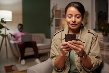 African young woman watching something on her mobile phone with man working on laptop in the background at office