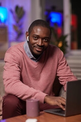 Portrait of African young man in headphones looking at camera while sitting on sofa in front of the table and typing on laptop