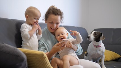 Young mother plays games on phone with two girl twins and curious pet on sofa. Home quality time of children and parents 