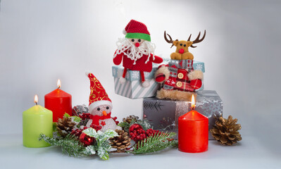 New year's composition: a toy Santa Claus, a reindeer and a Snowman, burning colored candles and gifts, a Christmas wreath with fir cones and red berries and gift boxes on a light background.