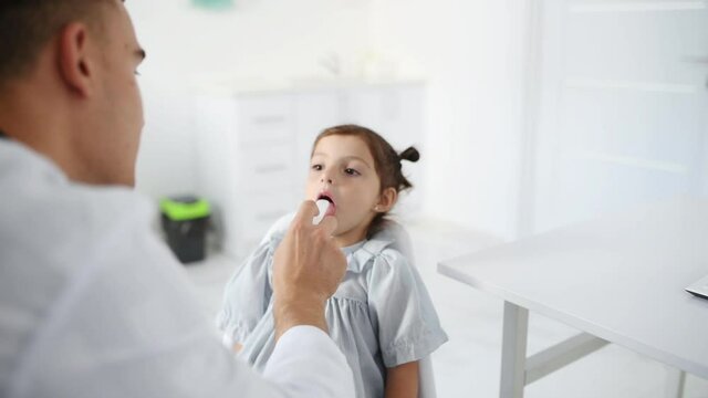 Young doctor in white coat cheking throat of little girl by using medical tonsil.