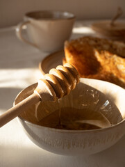 Composition of homemade thin pancakes on a beige ceramic plate with acacia honey in a jar and in a ceramic bowl with a wooden spoon and green tea in a beige ceramic cup on the table in the morning sun