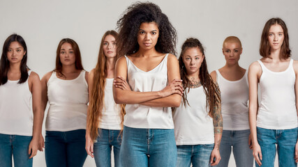 Portrait of young african american woman in white shirt and denim jeans posing with crossed arms. Group of diverse women standing isolated over grey background