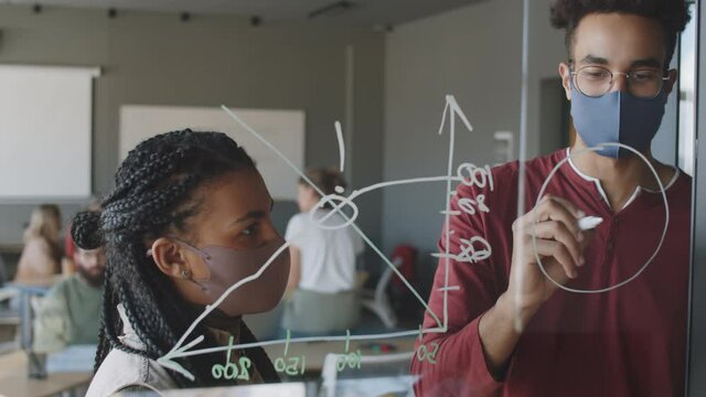 Two young male and female multiethnic colleagues in protective face masks drawing business chart on glass wall with markers while working together in office