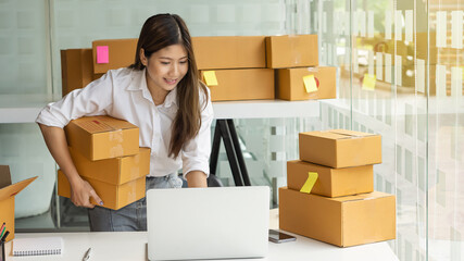 A beautiful Asian entrepreneur holds the boxes and checks the orders on her computer desktop.