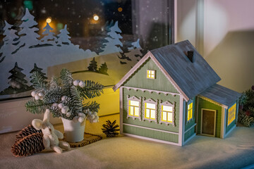 On windowsill, among Christmas tree decorations, night light in form of village house glows, against background of night snow window and paper decorations.