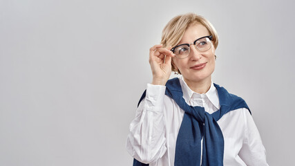 Portrait of elegant middle aged caucasian woman wearing business attire adjusting her glasses, smiling aside while standing isolated over grey background