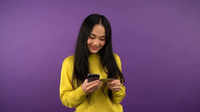 smiling young asian woman in bright yellow sweater is shopping online and paying with gold card via smartphone and shows tumb up 4K