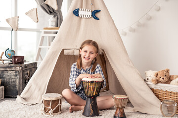 Little girl playing on traditional african djembe drums sitting in wigwam at home