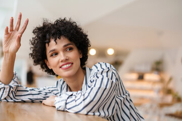 Happy curly woman smiling and waving hand while sitting in cafe